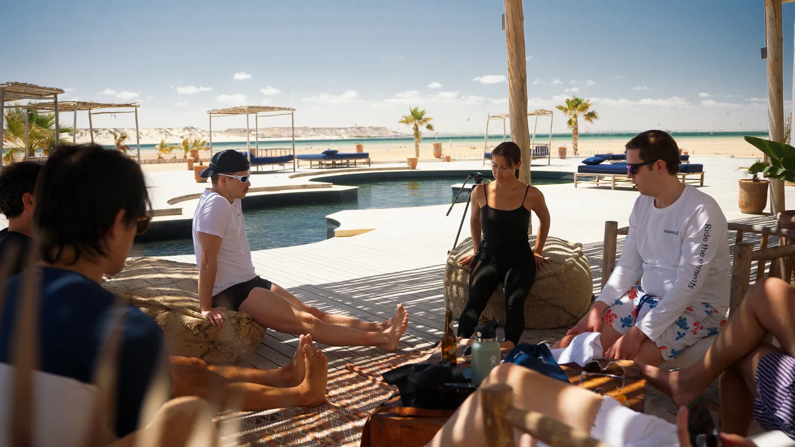 Group relaxing in a shaded lounge by the pool, with kites over the lagoon behind