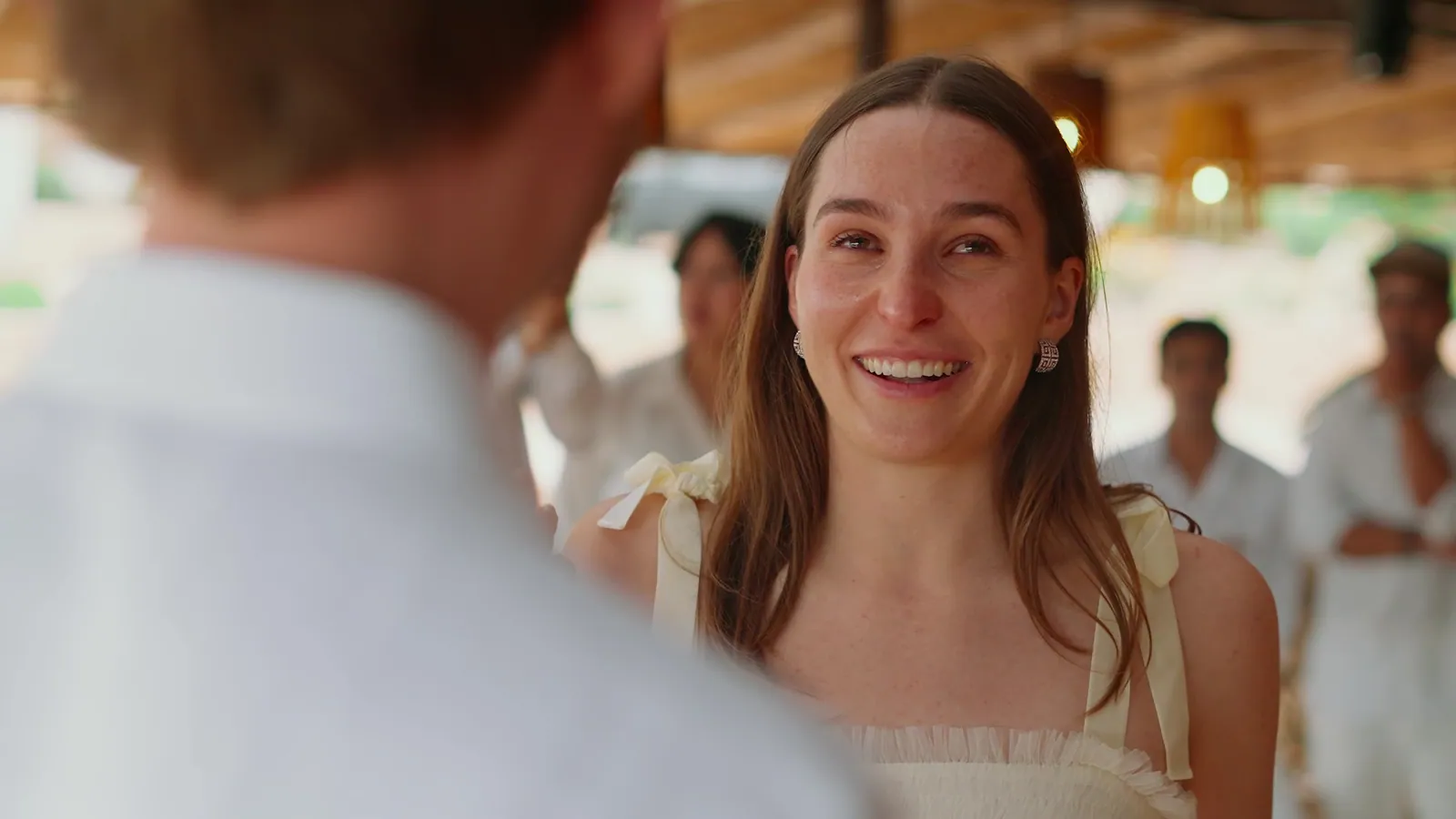 Bride smiling through tears during the vows, guests dressed in white behind her